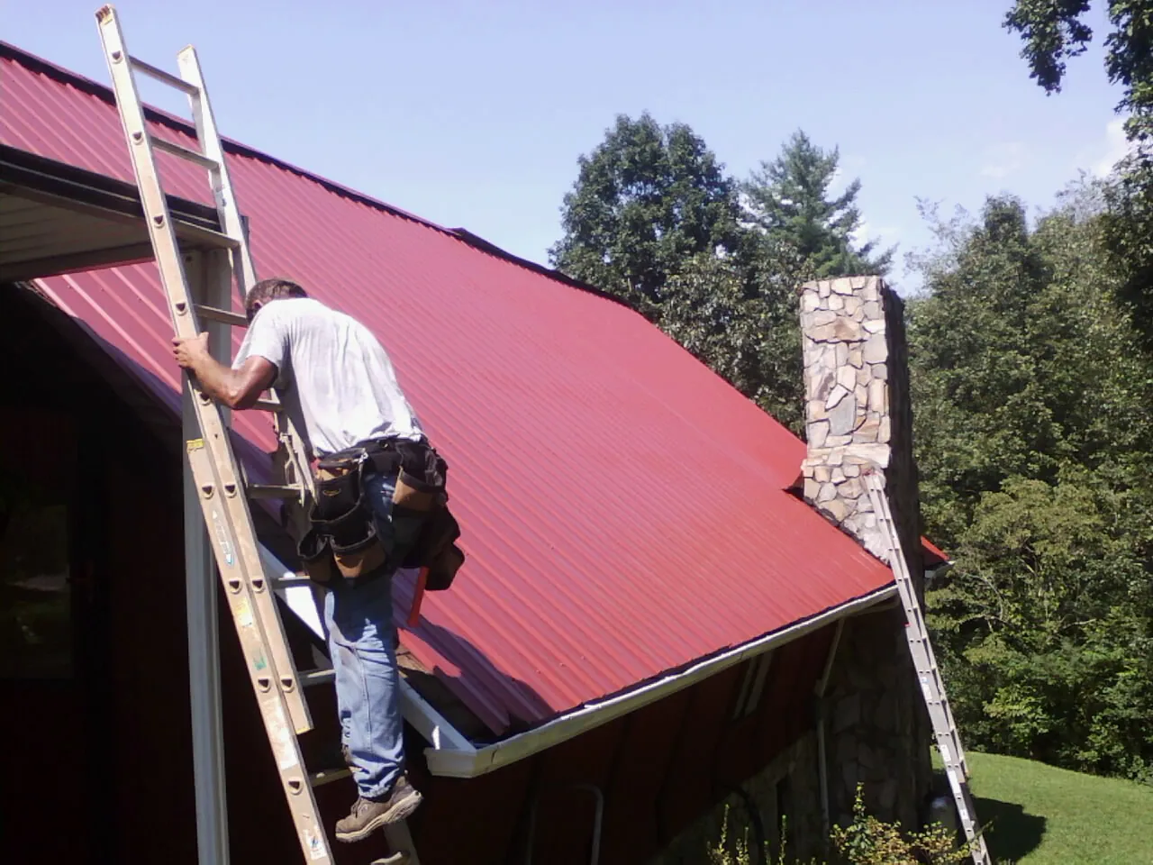 Roofer on a ladder beside a red metal roof and stone chimney — Soffit & Fascia Repair by Roofing Brothers in Potlatch